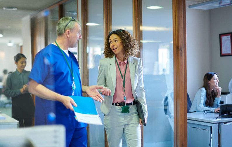 A healthcare consultant confers with a healthcare provider in a hospital hallway.